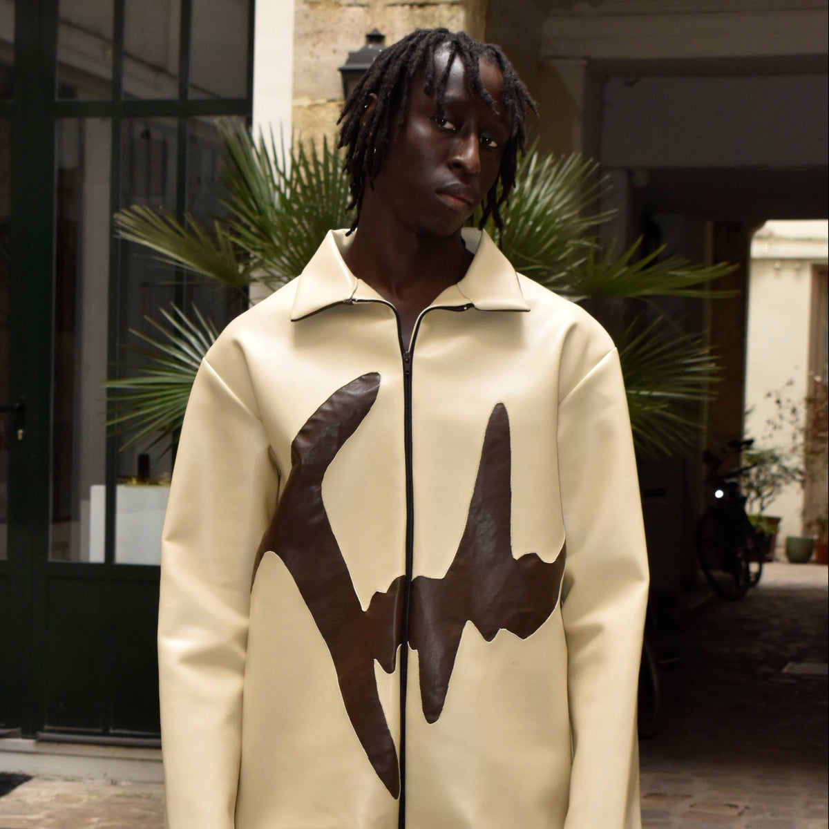 A man wearing a cream-colored zip-up jacket with a large dark brown abstract graphic print on the front, standing in an outdoor courtyard setting.